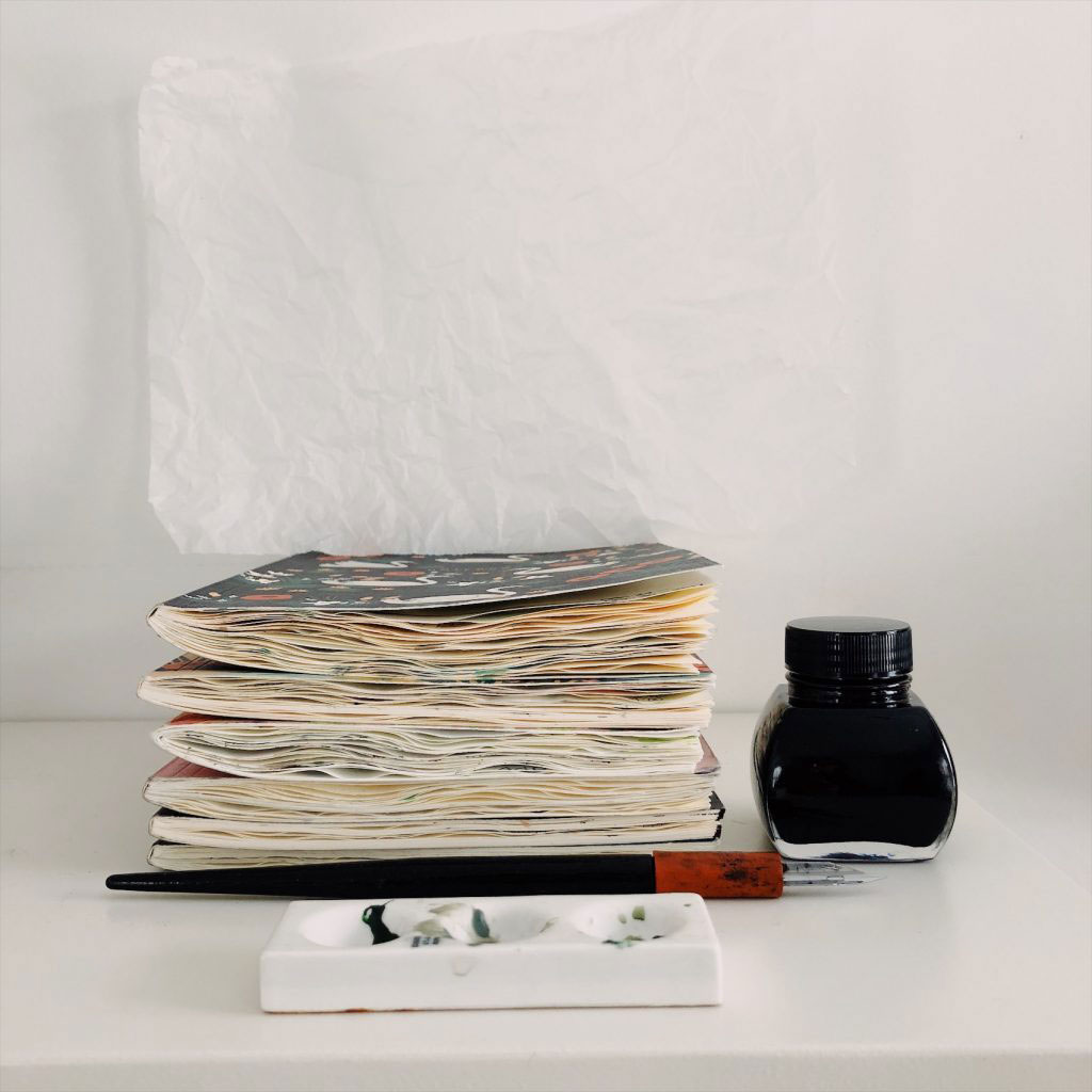 photo of a pile of full sketchbooks, ink and inkwell